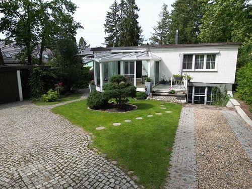 Weißes Haus mit Wintergarten und gepflegtem Garten, umgeben von Bäumen und zwei Zufahrten.