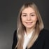 Junge blonde Frau in schwarzem Blazer und weißem Hemd lächelt in die Kamera vor grauem Hintergrund.
