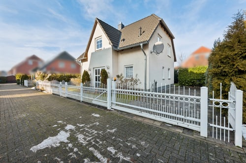 Haus mit weißem Metallzaun und gepflasterter Auffahrt, teilweise mit Schnee bedeckt, unter blauem Himmel.