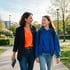 A woman and a young girl walk together on a paved park path, smiling and looking at each other.
