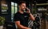 Ein lächelnder Mann im Sportivo-Shirt telefoniert im Fitnessstudio vor einem Laptop und Trainingsgeräten.