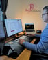 Mann mit Brille am Schreibtisch, arbeitet an Computer und Laptop, mit "RupCommerce" Logo an der Wand.