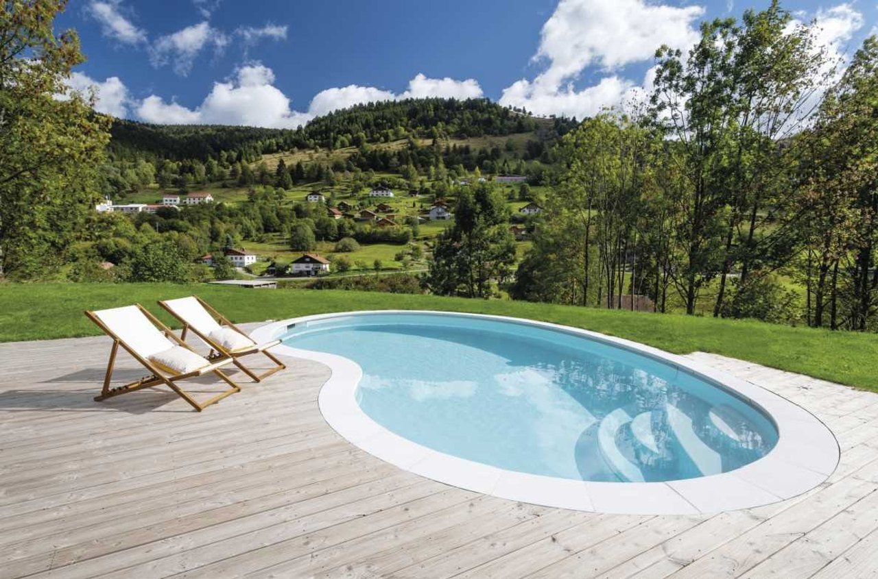 Nierenförmiger Pool auf einem Holzdeck mit zwei Liegestühlen, dahinter grüne Hügel und Häuser.