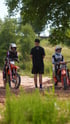 Zwei Motocross-Fahrer auf Motorrädern mit einem stehenden Mann dazwischen auf einer schlammigen Strecke.