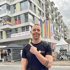 Ein lächelnder Mann zeigt auf ein Gebäude mit Regenbogenflaggen und einem Banner mit der Aufschrift „Das Haus nebenan“.