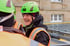 Ein lächelnder Arbeiter in leuchtend grünem Helm und orangefarbener Weste spricht mit einem Kollegen.