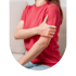 A person in a red t-shirt sits on a couch and grips their upper left arm with their right hand.