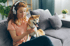 Rothaarige Frau mit Kopfhörern sitzt auf einem grauen Sofa und zeigt ihrem Shiba Inu Hund ein Smartphone.