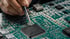 A hand uses a pointed tool to work on a black microchip embedded in a green circuit board.