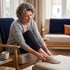 An older woman sits in a wooden armchair and holds her ankle while resting her foot on a cushioned ottoman.