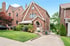 A red brick house with a steep gable roof, awning, and arched doorway, next to a driveway and green lawn.