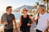 Drei aktive Senioren in Sportkleidung spazieren lachend mit Yogamatte und Wasserflasche an einer sonnigen Strandpromenade.