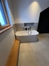 Eine weiße Badewanne steht neben einer Holzsitzbank in einem modernen Badezimmer mit grauen Fliesen Reutte