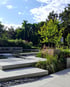 Moderner Garten mit grauen Steinstufen, Wasserbecken, Pflanzen und Bäumen unter blauem Himmel.