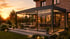 Moderner Kaltwintergarten aus Glas mit Beleuchtung und Gartenmöbeln vor einem Backsteinhaus bei Sonnenuntergang.