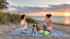 Zwei Frauen und ein Border Collie meditieren im Sonnenuntergang auf einer Decke am Strand.