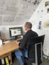 Ein Mann sitzt an einem Schreibtisch und arbeitet an einem Computer mit 3D-Modellierungssoftware, umgeben von technischen Zeichnungen.