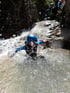Person in Neoprenanzug und blauem Helm rutscht eine felsige Wasserrutsche hinunter, Wasser spritzt.
