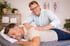 Therapeut massiert Rücken eines lächelnden Patienten auf einem Massagetisch in einer Klinik.