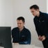 Zwei Männer in blauen Pullovern schauen zusammen auf einen Computerbildschirm.