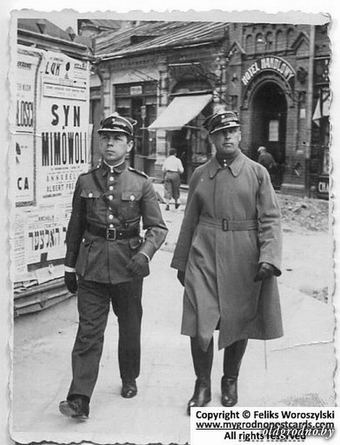 Después del pogromo. Foto de julio de 1935 de la misma esquina Dominikańska × Elizy Orzeszkowej en Grodno, un mes después del pogromo antisemita del 7 de junio. Dos policías de la Policja Państwowa polaca caminan por la vereda — uno de uniforme oscuro completo, cinturón cruzado, casco; el otro con sobretodo gris largo, guantes y gorra. A la derecha de la imagen, donde camina el oficial del sobretodo, se ven claramente escombros, ladrillos rotos y material de obra desparramados en la calle. Al fondo se distingue el Hotel Handlowy con su rótulo y el panel de afiches del mismo cine Łuk —pero con películas distintas en cartelera respecto a la foto anterior—.