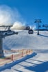 Schneekanonen erzeugen Schnee auf einer Skipiste mit Gondeln, die über eine Absperrung fahren.