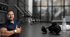 Muskulöser Mann lächelt und gibt einen Daumen hoch neben Hanteln und Wasserflasche im Fitnessstudio.