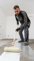 Mann im Arbeitsanzug mit Handschuhen rollt weiße Grundierung auf einen Betonboden.