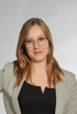 Eine junge Frau mit braunen Haaren, Brille, schwarzem Oberteil und grauem Blazer lächelt.