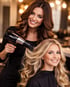 Friseurin föhnt einer lächelnden Kundin mit langen blonden Locken die Haare in einem Salon.