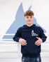 Junger Boxer im dunkelblauen Hoodie mit Aufschrift „WE LOVE BOXING“ lehnt lächelnd an einem weißen Boxring.