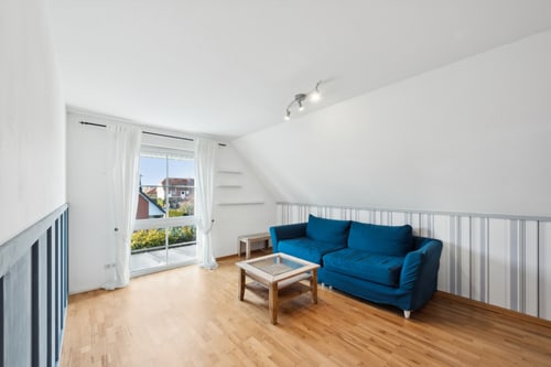 Heller Wohnraum mit schrägem Dach, blauer Couch, Couchtisch und Fenster mit Blick ins Grüne.