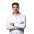 A man in a white button-down shirt with black buttons stands with arms crossed against a black background.
