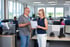 Zwei Kollegen besprechen Dokumente in einem Großraumbüro mit Computern und Fenstern.