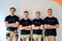 Vier lächelnde Handwerker in schwarzen T-Shirts und beiger Arbeitshose stehen vor einer weißen Wand.
