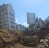Bagger gräbt eine Baugrube vor einem weißen Gebäude mit Gerüst und einer Schallschutzwand.