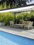 Terrasse mit Holzmöbeln und weißer Pergola neben einem Pool, umgeben von grünen Büschen.