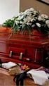 Дерев'яна труна з ручками, прикрашена білими квітами та зеленню, стоїть на коричневій поверхні.