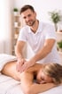 Ein lächelnder Masseur massiert einer blonden Frau den Rücken, die entspannt auf einem Handtuch liegt.