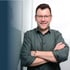 Lächelnder Mann mit Brille und dunkelgrünem Hemd steht mit verschränkten Armen vor weißem Hintergrund.