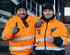 Zwei lächelnde Männer in orangefarbenen Arbeitsjacken und Mützen zeigen Daumen hoch.