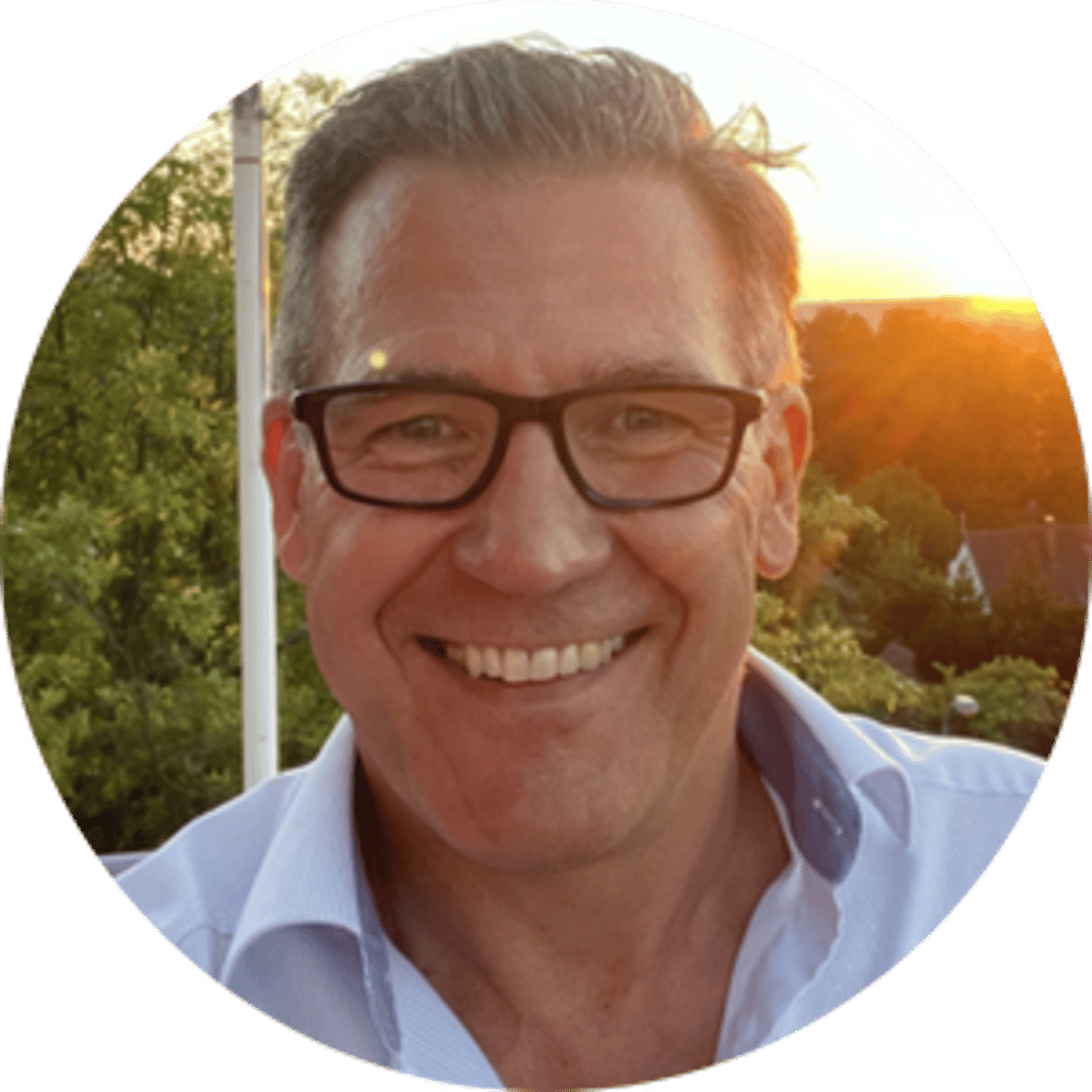 Ein lächelnder Mann mit Brille und hellblauem Hemd vor Bäumen und Sonnenuntergang. Frank Freitag