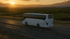Weißer Reisebus fährt bei Sonnenuntergang auf einer Autobahn durch eine ländliche Landschaft.