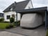 Ein weißes Wohnhaus mit Carport, darunter ein graues Wohnmobil unter einer Schutzhülle auf einem gepflasterten Hof.