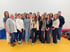 Eine Gruppe von 18 Erwachsenen, meist Frauen, steht in einem Raum mit gelbem Boden und weißer Wand.