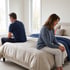 A man and woman sit on opposite sides of a bed facing away from each other in a bright bedroom.
