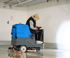 Ein Mann in einer Garage fährt eine blaue Aufsitz-Bodenreinigungsmaschine und reinigt den glänzenden Betonboden.