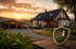 Modernes Wohnhaus und Scheune bei Sonnenuntergang mit einem Wappen-Logo für landwirtschaftliche Versicherungen im Vordergrund.