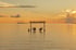 Zwei Personen sitzen bei Sonnenuntergang auf mondförmigen Schaukeln im flachen Wasser unter einem Schild „Visit Dhangethi“, eine dritte Person steht daneben, am Horizont eine kleine Insel