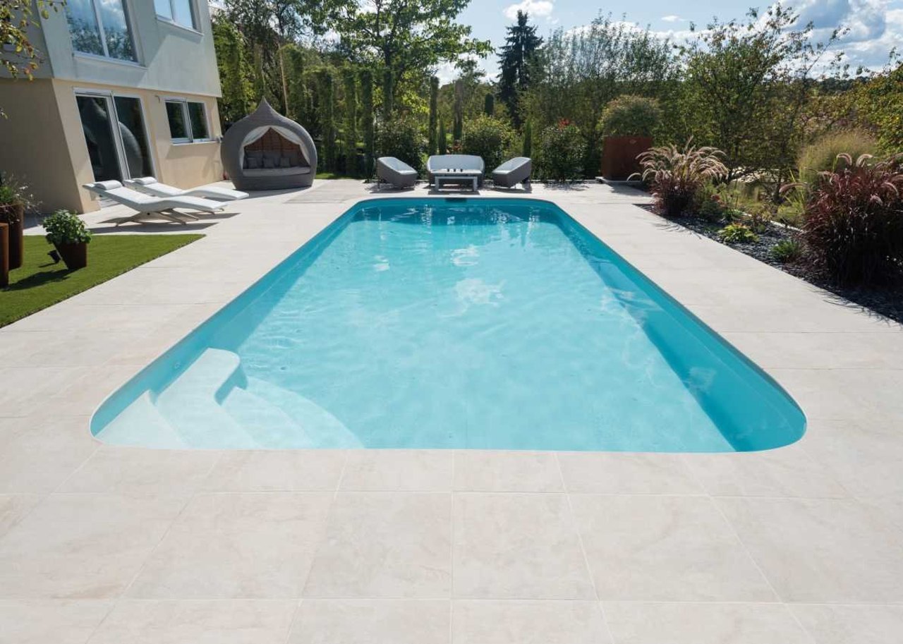 Blauer Swimmingpool mit Stufen im Garten, dahinter ein Haus und Liegestühle.